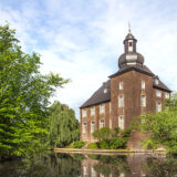 Historisches Wasserschloss mit Turm und Backsteinfassade, umgeben von Bäumen und einer spiegelnden Wasserfläche an einem sonnigen Tag.